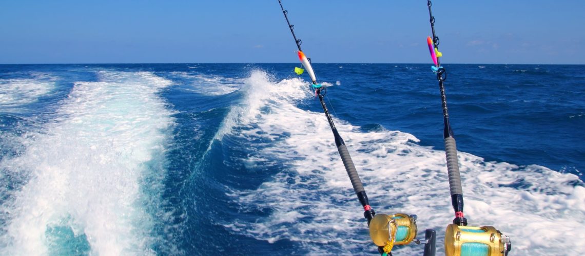 Two fishing rods are mounted on the back of a moving boat, with the ocean and waves visible under a clear blue sky.